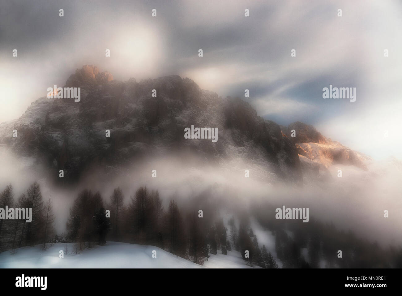 Fog around the mountain Sella with last rays of Sun on the top in winter season and woods in foreground, Dolomites - Italy Stock Photo