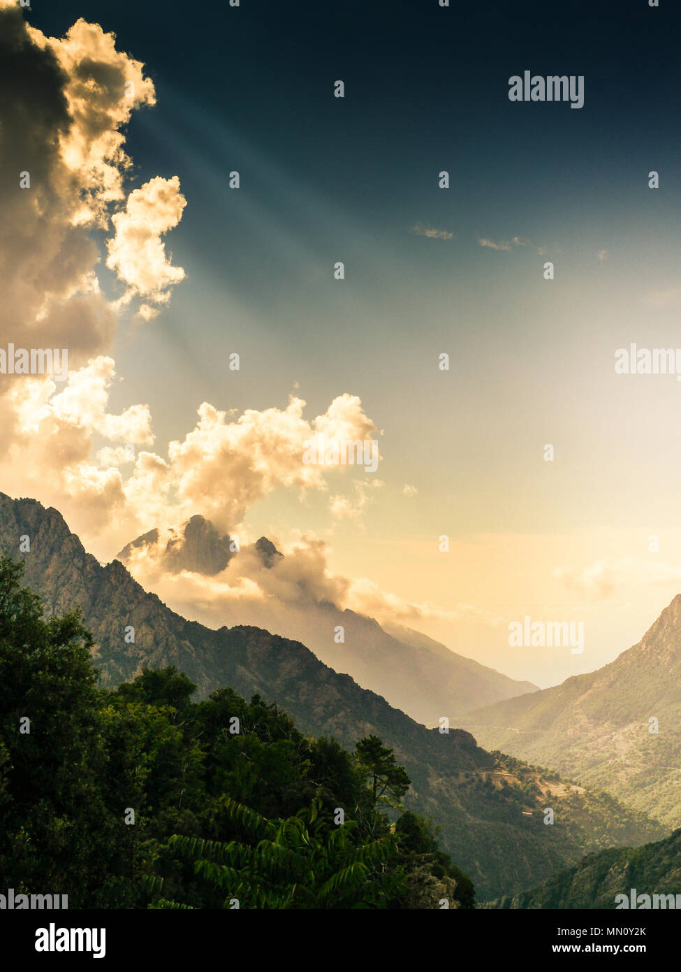 Mountainpeak surrounded by clouds and godrays with trees in the foreground on corsica. Misty sky Stock Photo