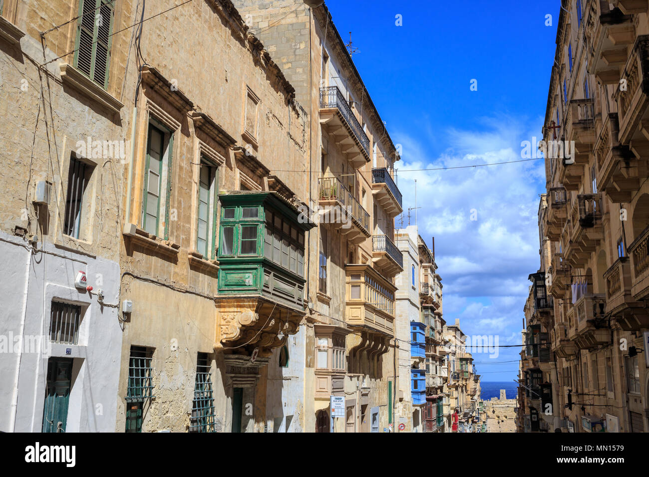 Malta, Valletta, traditional sandstone buildings with colorful wooden windows on covered balconies. Blue sky with clouds and sea background. Stock Photo
