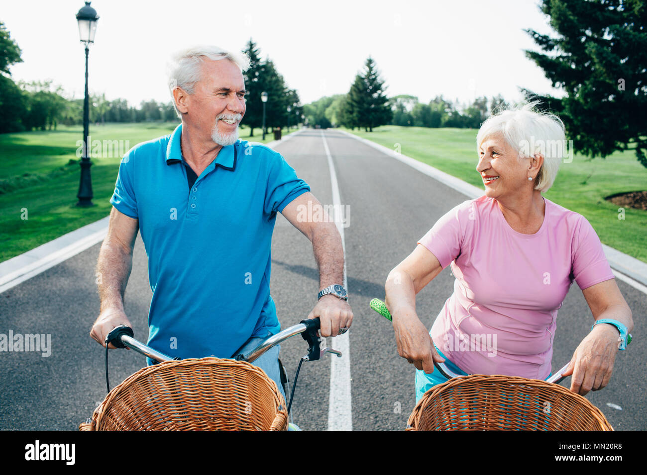 Older couple riding bicycles with baskets, standing in the middle of the road and looking into each other's eyes Stock Photo