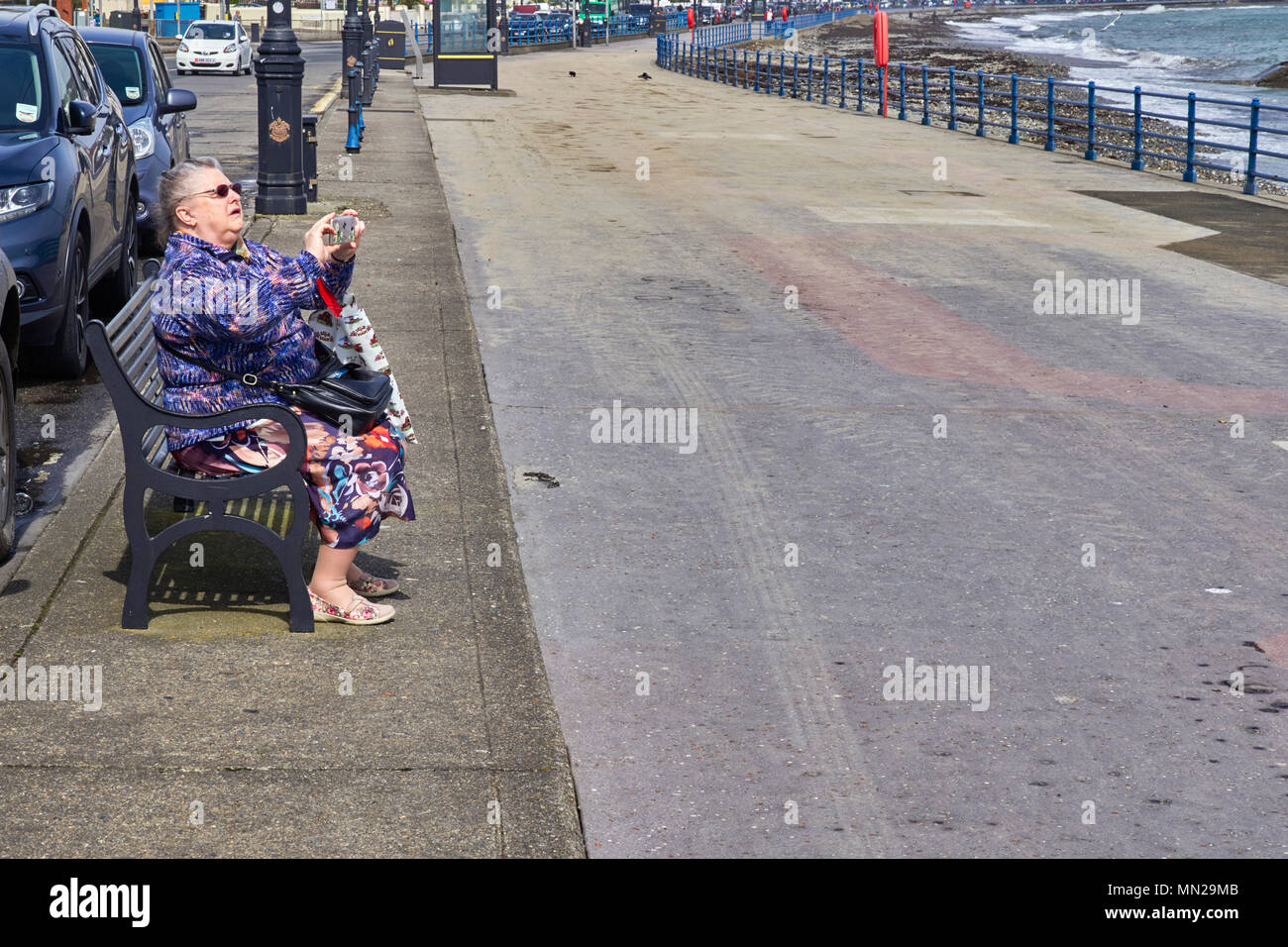 Older woman sitting on a seaside bench taking a photo with phone camera Stock Photo