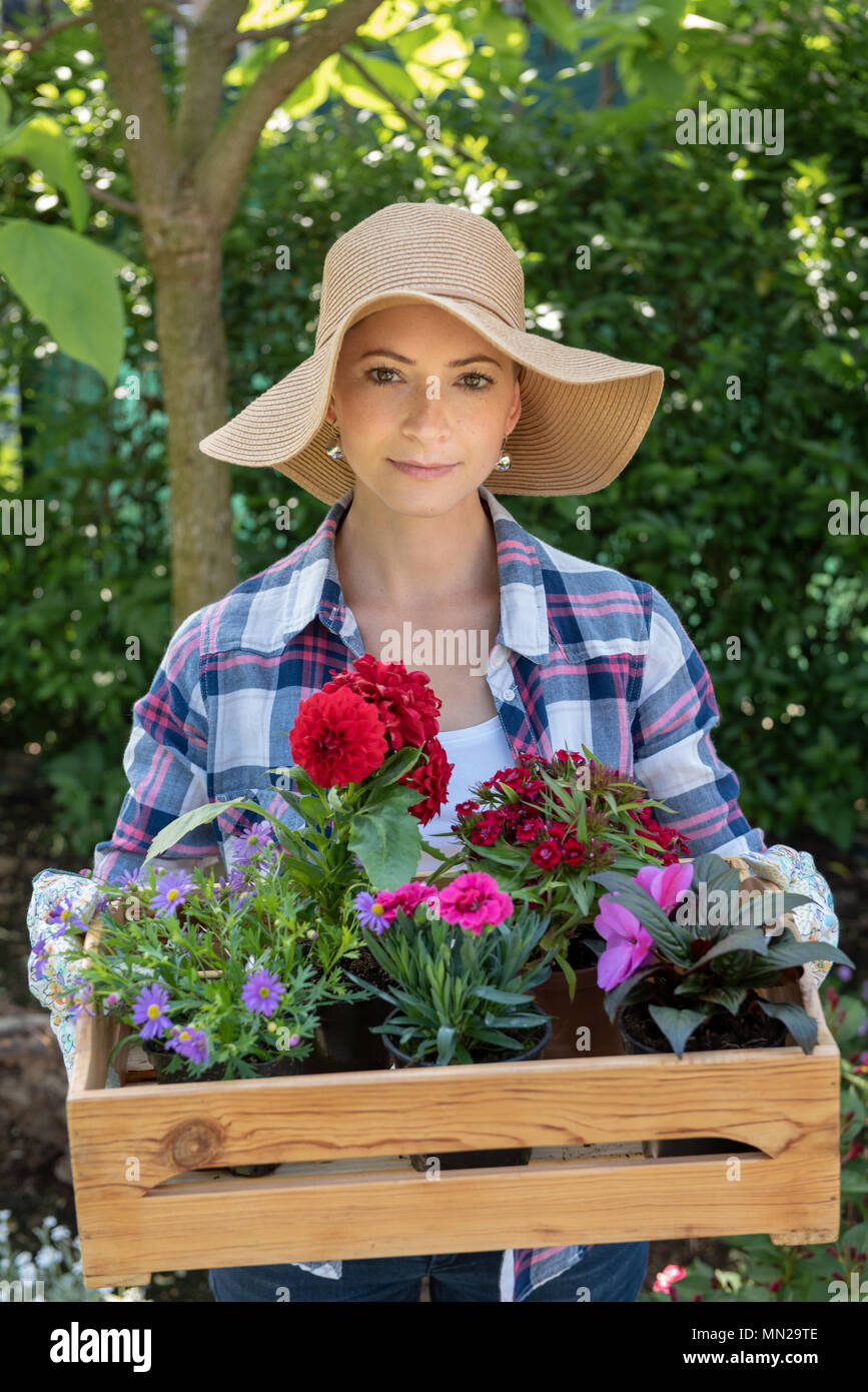 Beautiful female gardener wearing straw hat holding wooden crate full of flowers ready to be planted in her garden. Gardening concept. Stock Photo