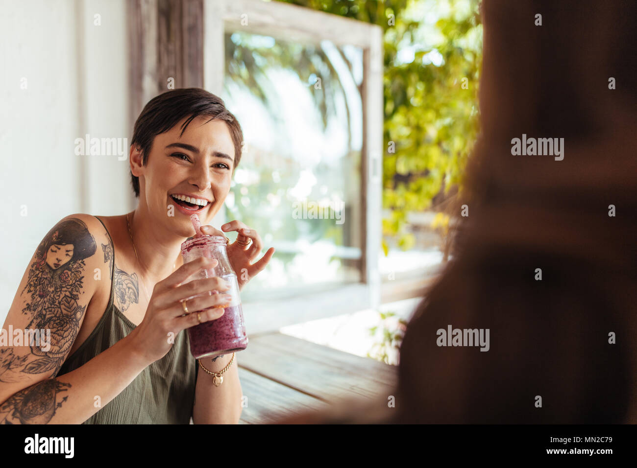 Woman holding a smoothie jar talking to her friend at a restaurant. Woman with tattoo on hand at a restaurant drinking juice. Stock Photo