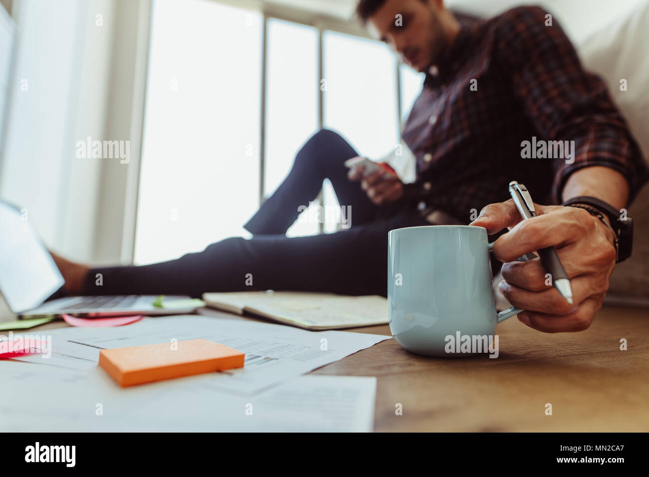Man working on laptop computer sitting on the floor at home. Freelancer sitting on floor looking at his mobile phone holding a coffee cup. Stock Photo