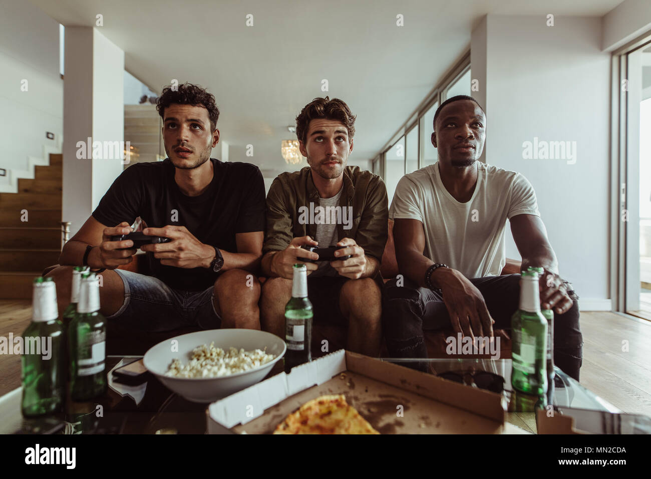 Friends having fun with drinks and snacks while playing video game at home. Two men playing video game holding joysticks while another man watches hol Stock Photo