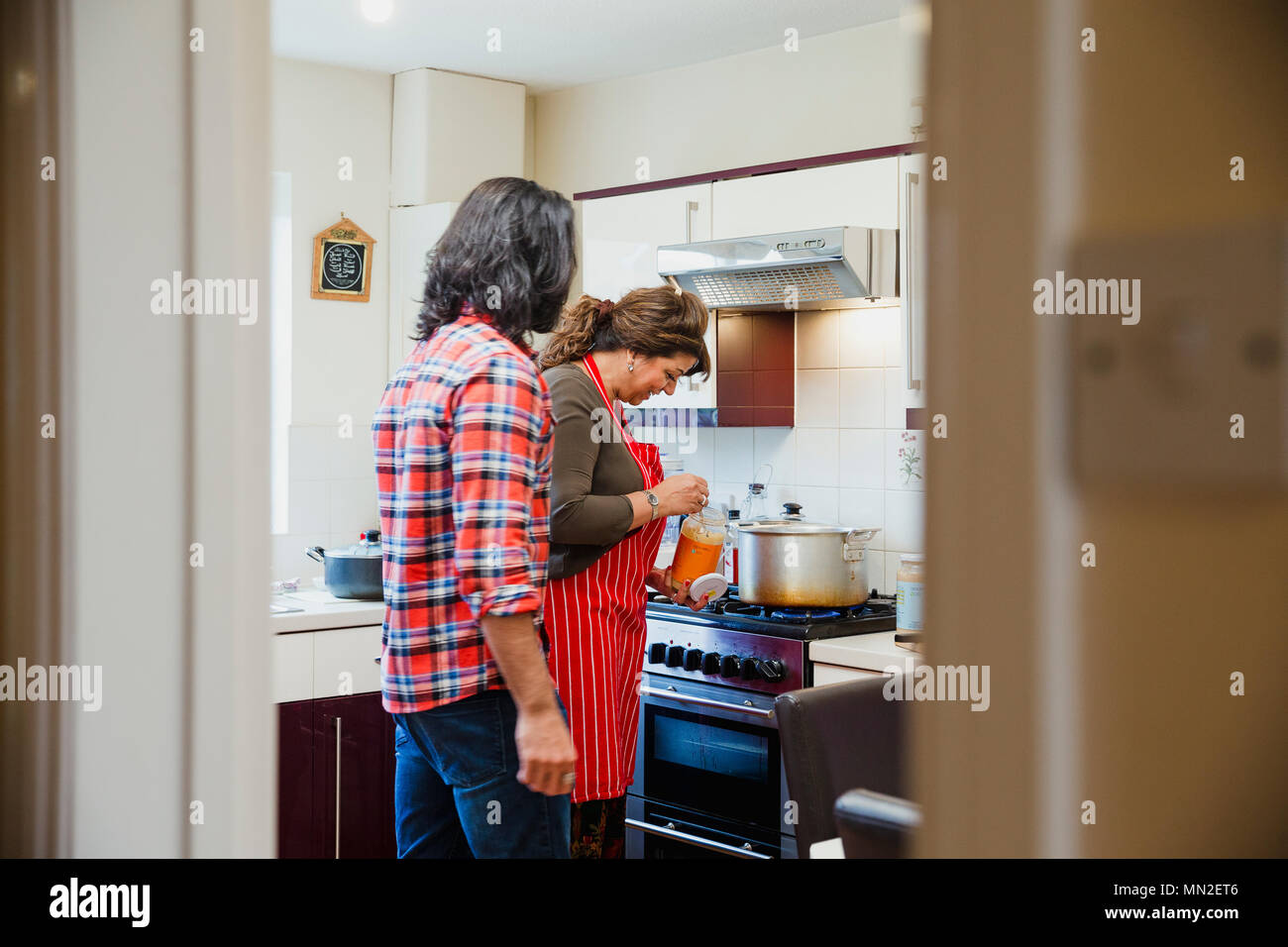 Mid adult man is relaxing in the kitchen talking to his mother while she prepares a curry on the cooker. Stock Photo