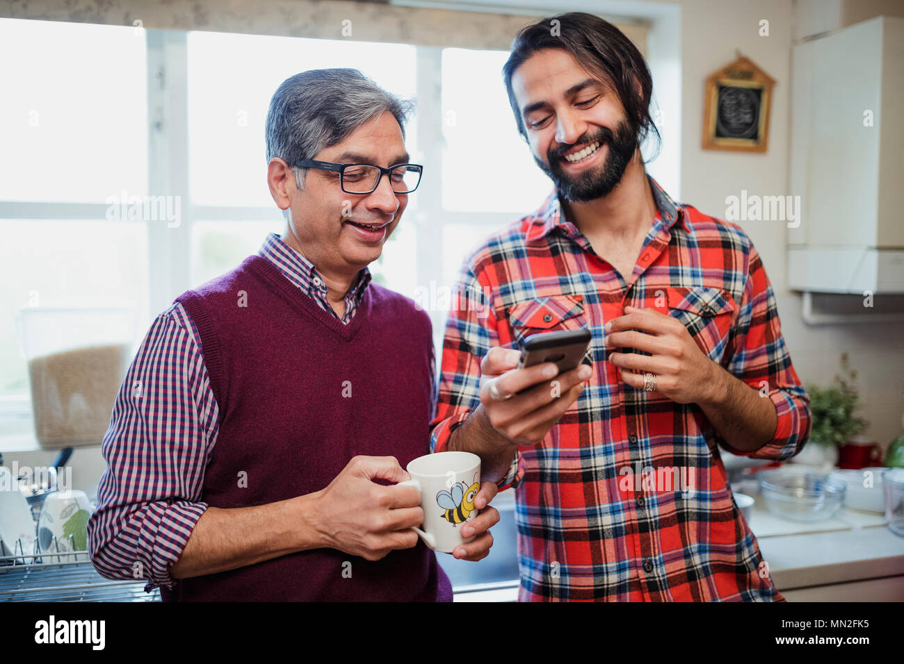 Mid adult man is showing his mature father something on his smart phone. He is holding a mug of tea. Stock Photo