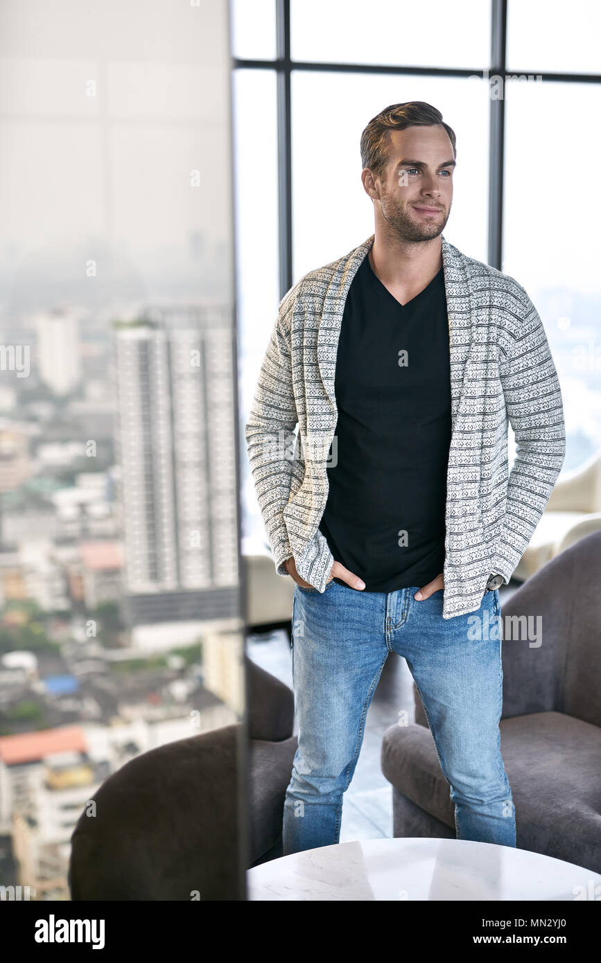 Handsome guy stands in his flat on the large windows background. He wears a  light gray cardigan, black T-shirt and blue jeans. Man holds his hands in  Stock Photo - Alamy