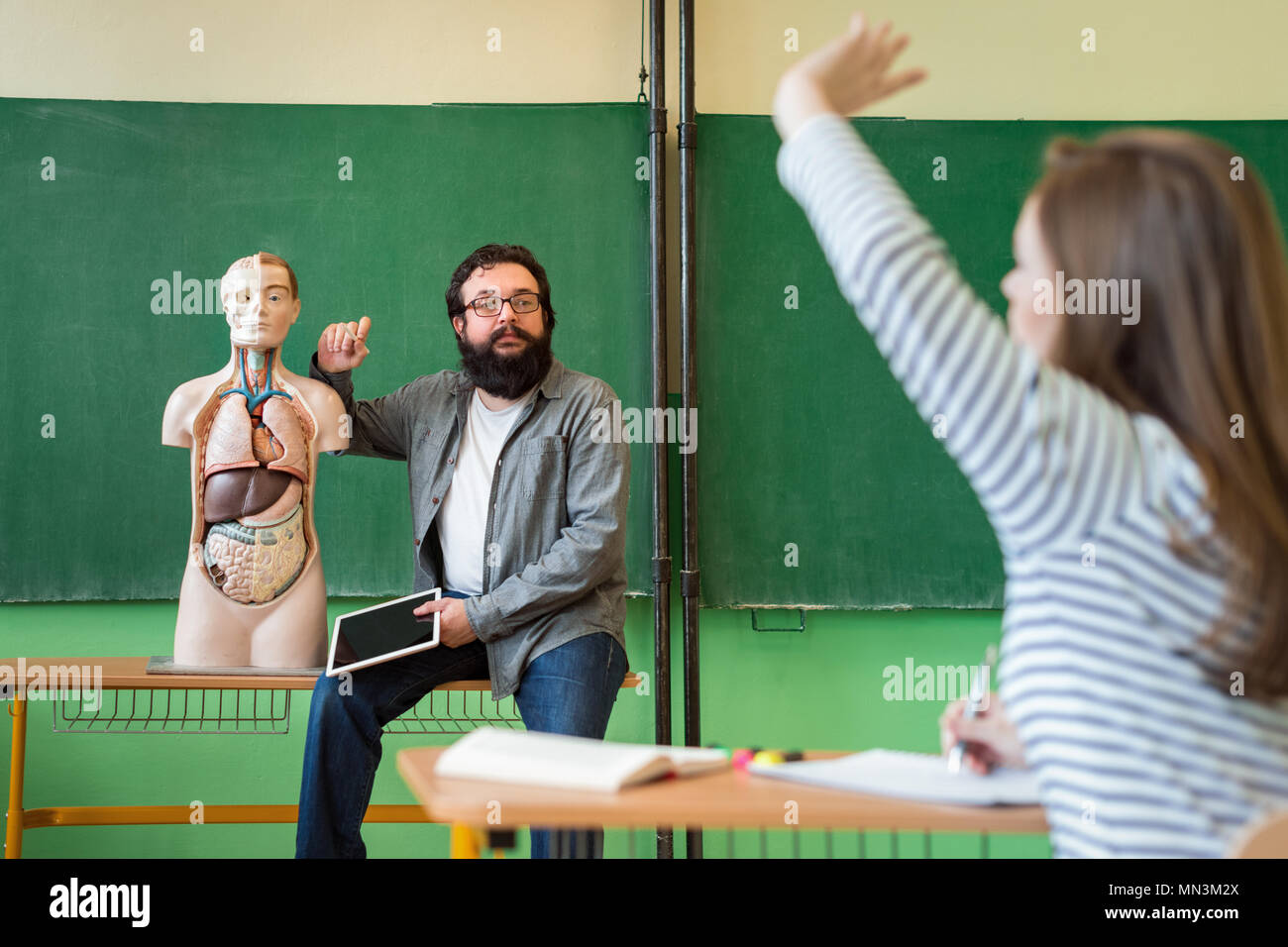 Young male hispanic teacher in biology class, holding digital tablet and teaching human body anatomy, using artificial body model. Stock Photo