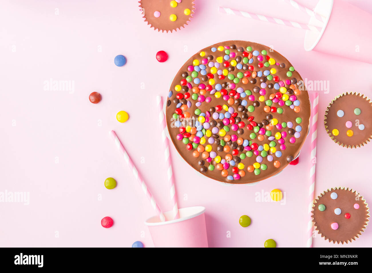 Milk Chocolate Birthday Cake with Multicolored Glazed Candy Sprinkles Buttercups Paper Drinking Cups Pink Straws Pink Background. Party Celebration Ki Stock Photo