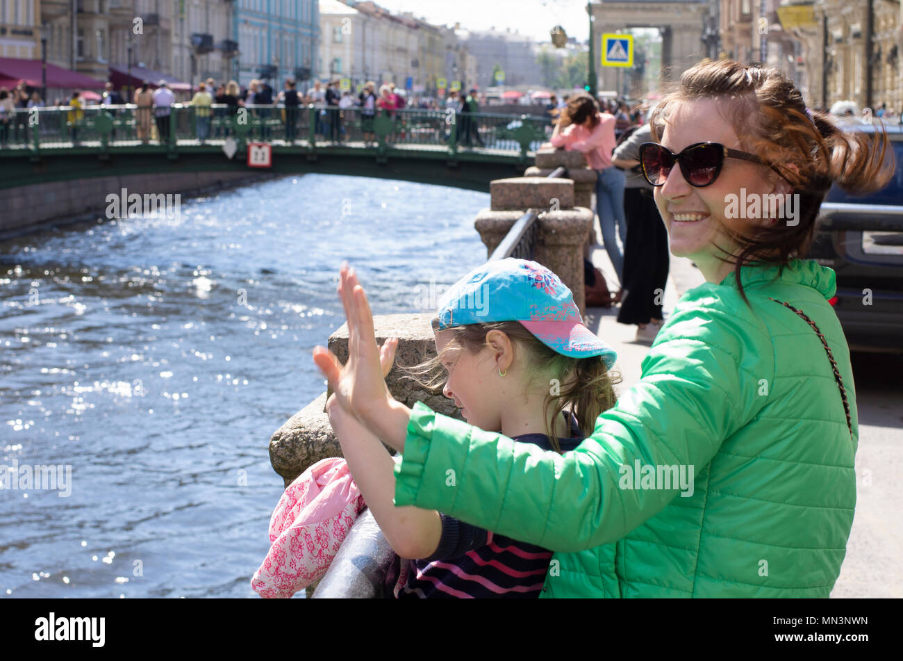 Young mother with the daughter smile and welcome tourists in sunny summer and windy day. On a blurred background Griboyedov canal in St. Petersburg Stock Photo