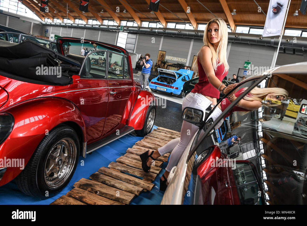 09 May 2018, Friedrichshafen, Germany: Miss Tuning Vanessa Schmitt leans  against a tuned VW Polo before the start of the fair "Tuning World  Bodensee". To the left is a tuned VW Beetle.