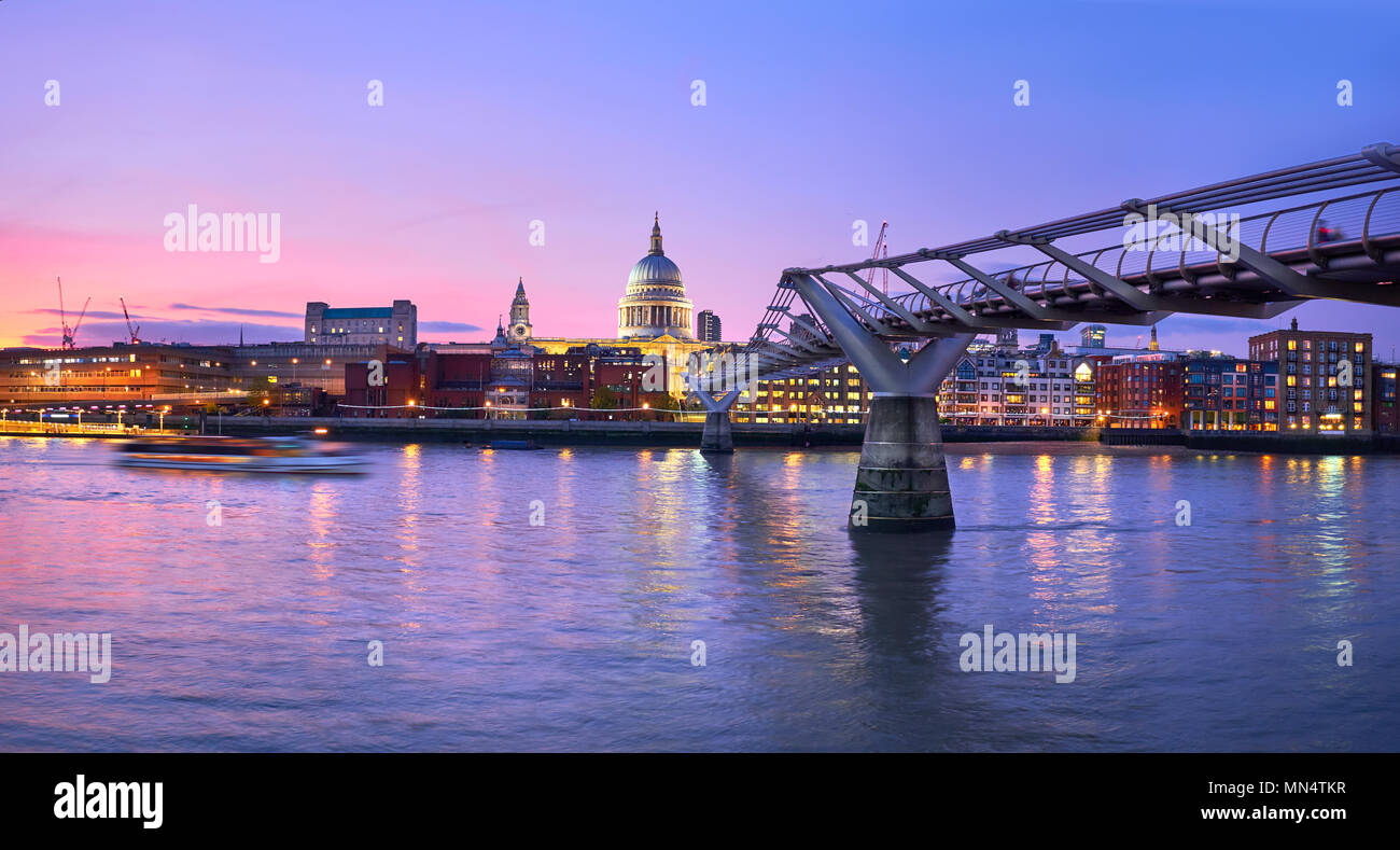 London at sunset, Millennium bridge leading towards illuminated St. Paul cathedral over Thames river with city bathing in electric light. Panoramic to Stock Photo
