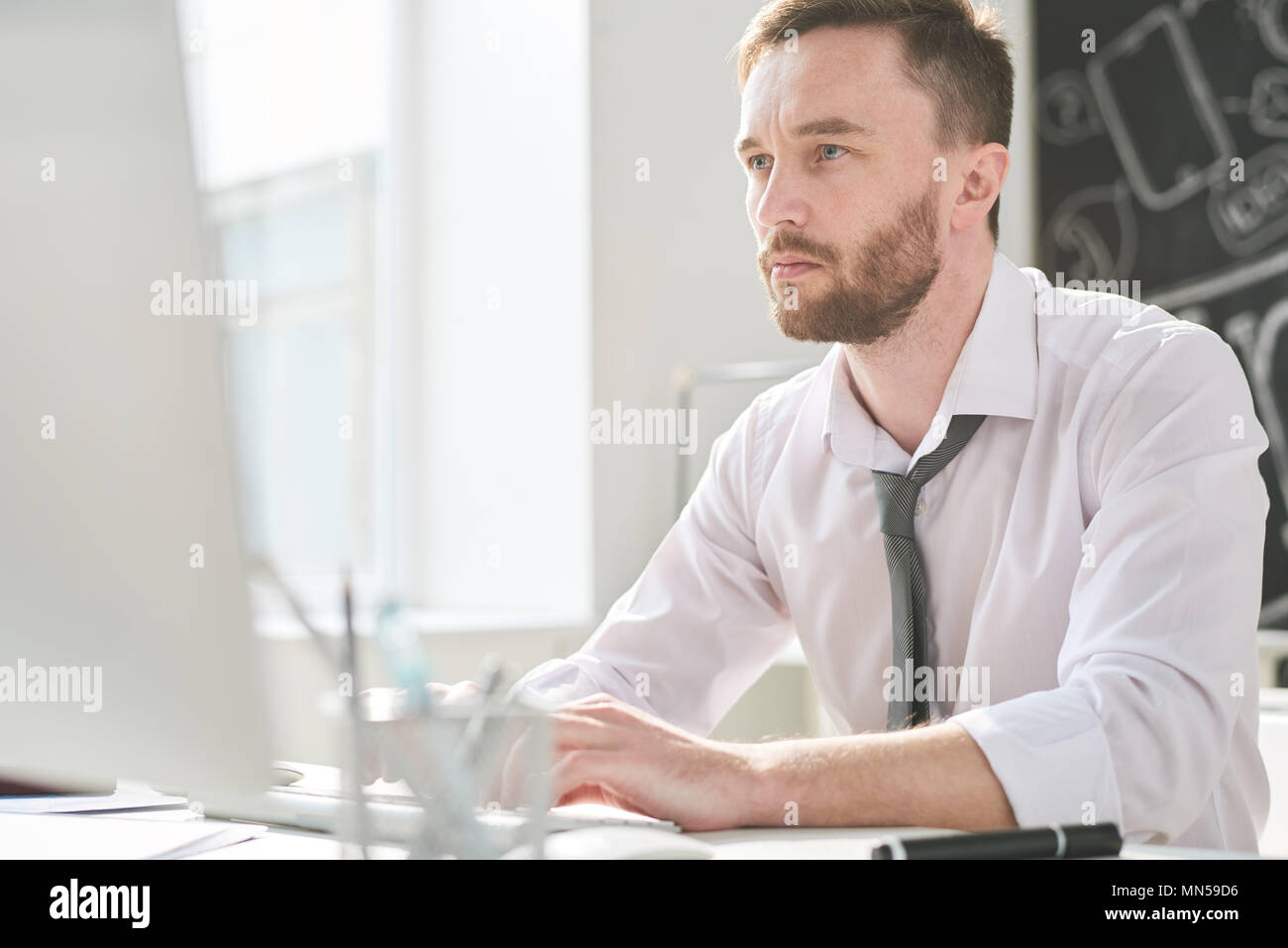 Portrait of thoughtful handsome businessman sitting at desk in office and using modern computer for work, copy space Stock Photo