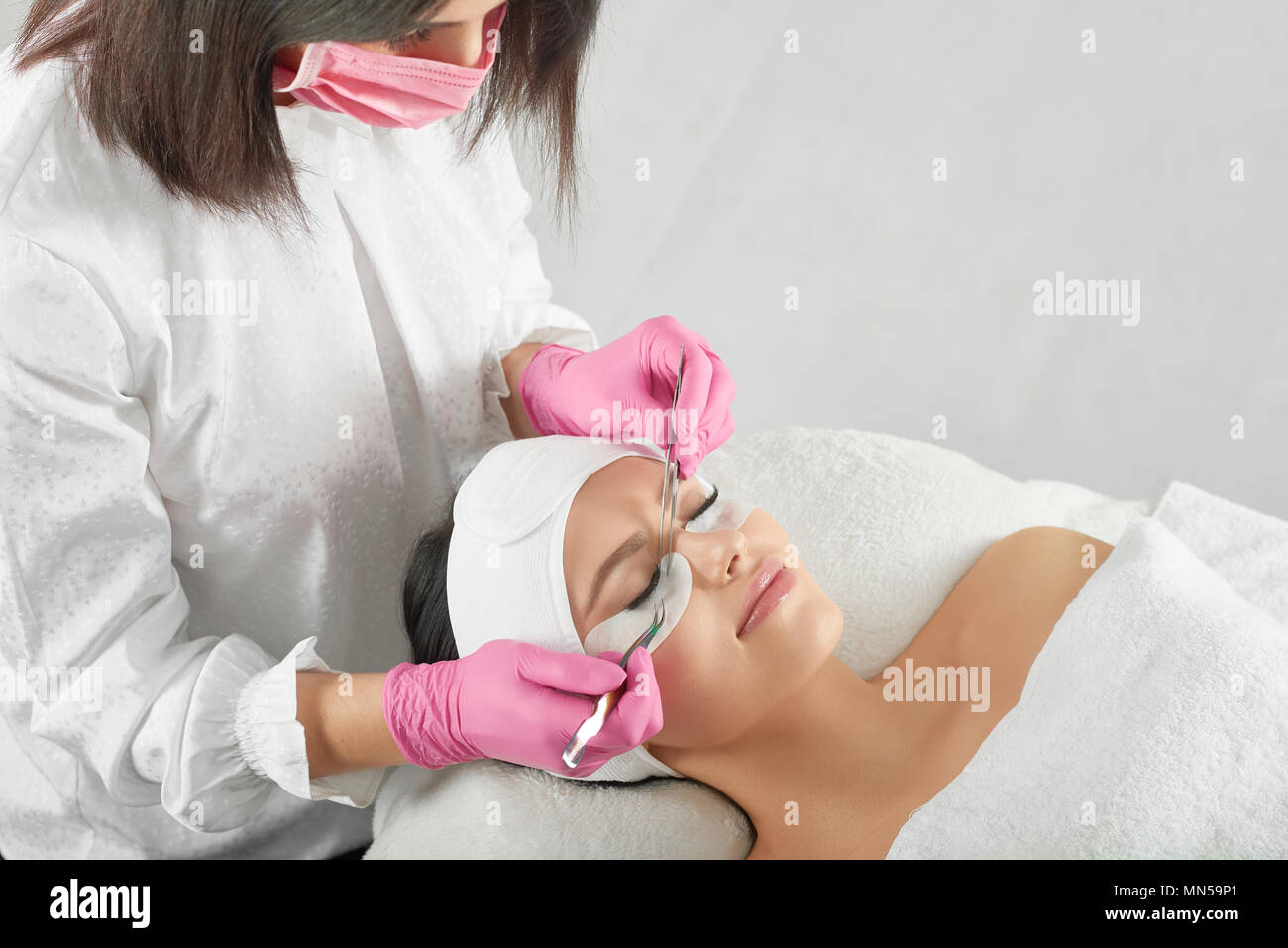 Professional lashmaker making long beatiful lashes. Woman lying on cosmetologiacal coach during lashes enlarging. woman with pretty face wearing headbandage and stickers for lashmaking . Stock Photo