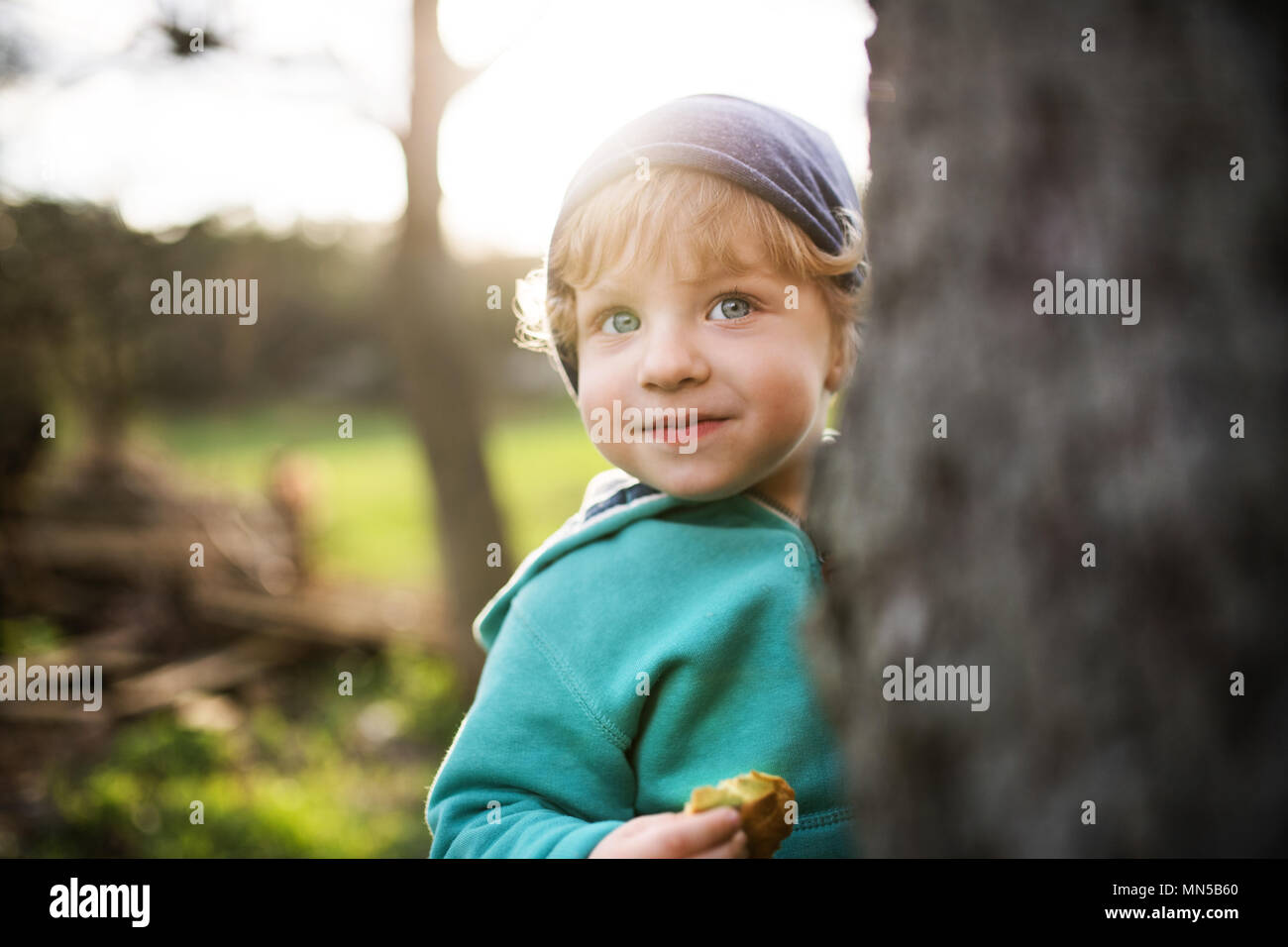 A happy toddler boy hiding behind the tree outside in spring nature. Stock Photo