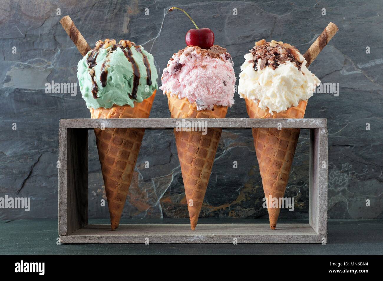 Pistachio, cherry and vanilla ice cream with topping in waffle cones in rustic wood holder over a slate background Stock Photo