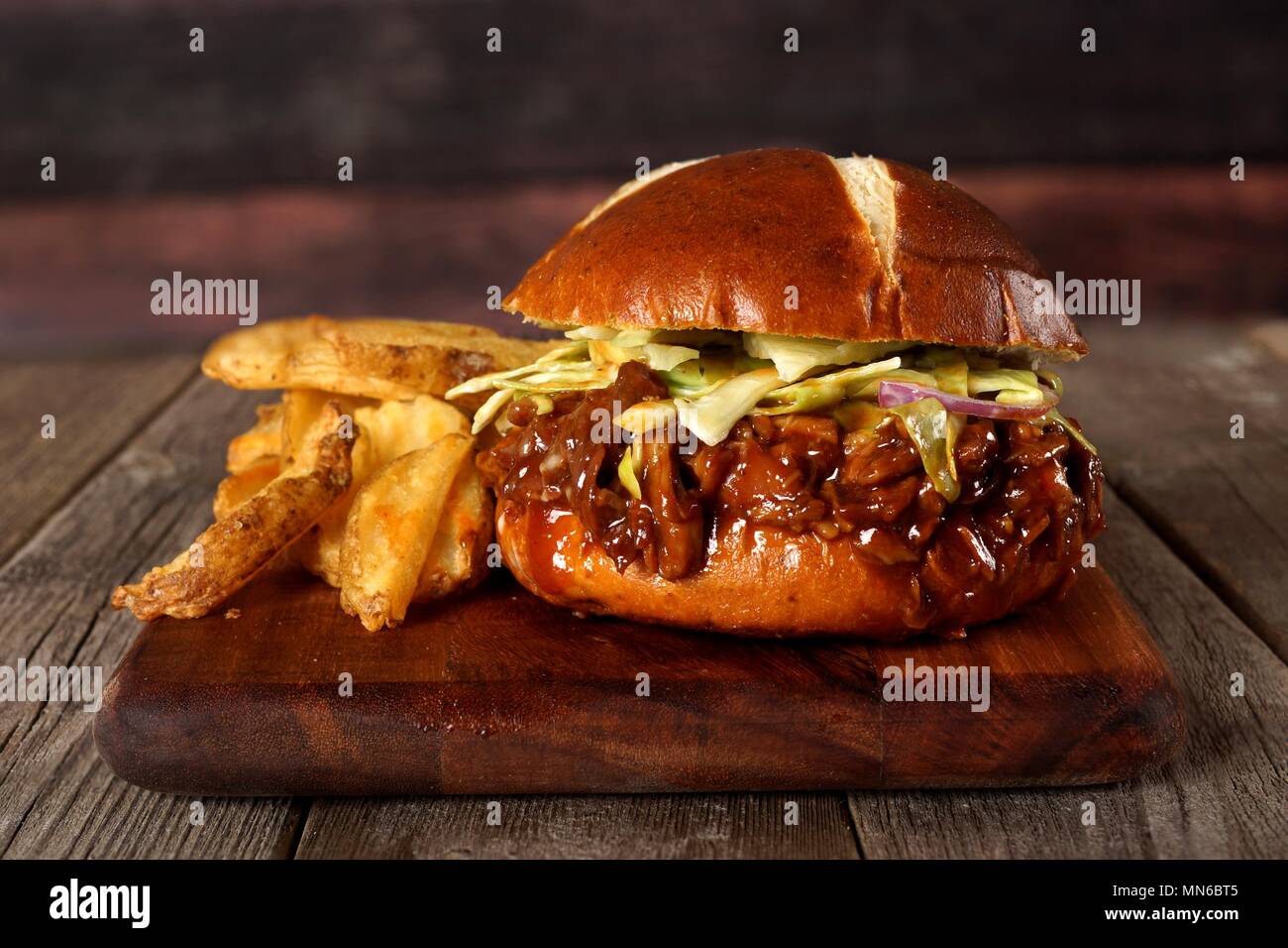 Pulled pork sandwich on pretzel bun with potato wedges on a serving board with wood background Stock Photo
