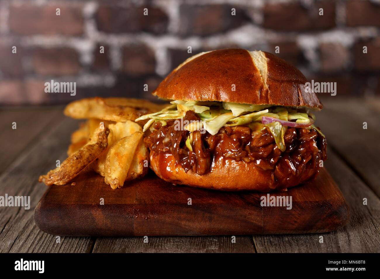 Pulled pork burger on pretzel bun with potato wedges on a wood server with brick background Stock Photo
