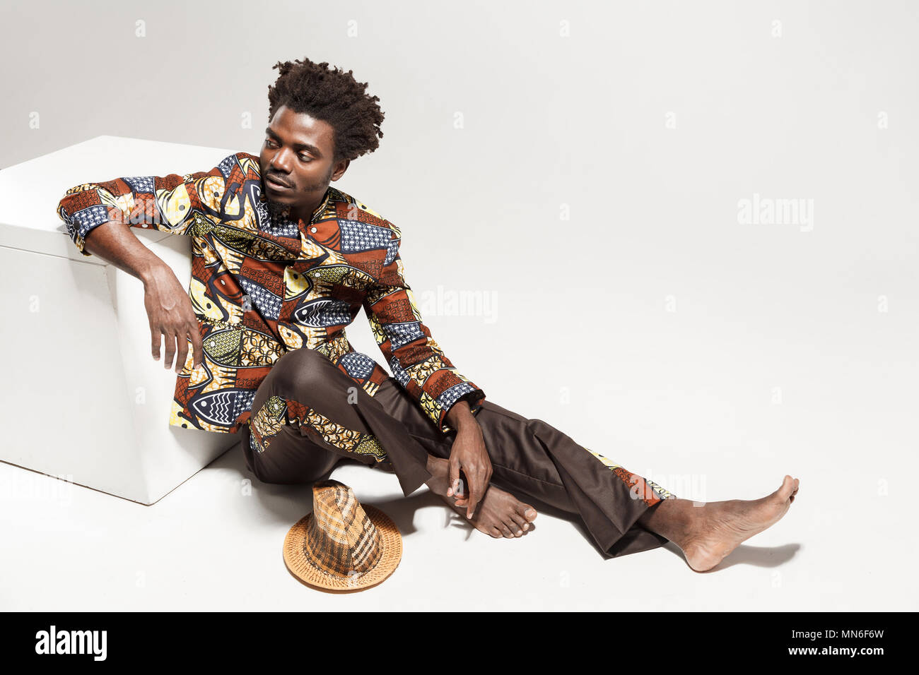 Wondering afro man in traditional national clothes sit on coub. Indoor, isolated on gray background Stock Photo