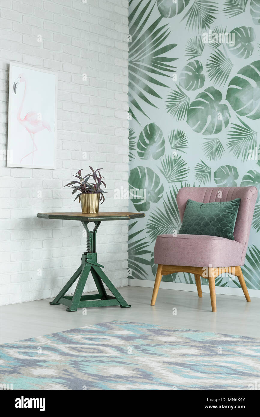 Plant in gold pot on green table next to pink chair with pillow in floral room with white poster Stock Photo