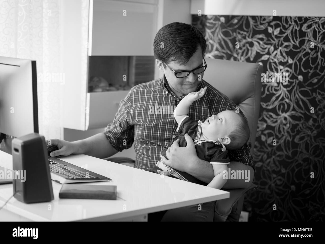Black and white image of smiling young man working in home office and looking after his baby son Stock Photo
