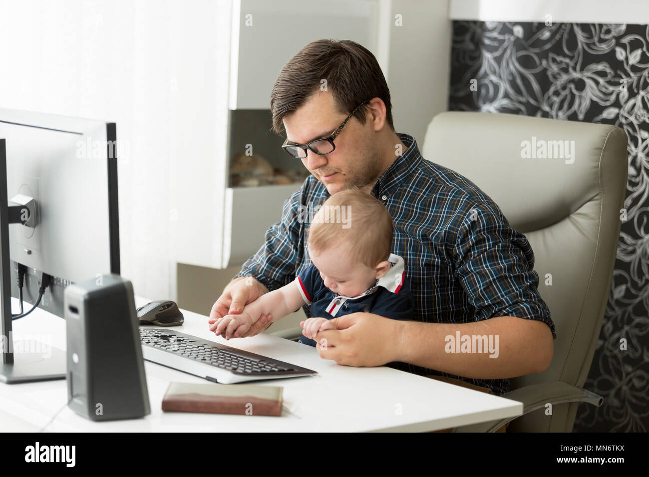 Portrait of young self-employed man working at home office on computer and looking after his baby son Stock Photo