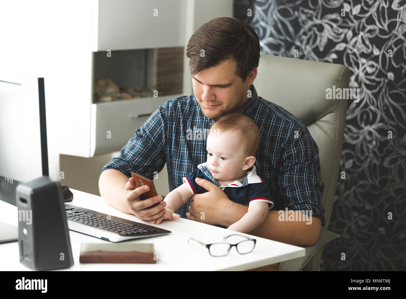 Toned portrait of young selfemployed man working at home with computer and mobile phone and looking after his baby son Stock Photo