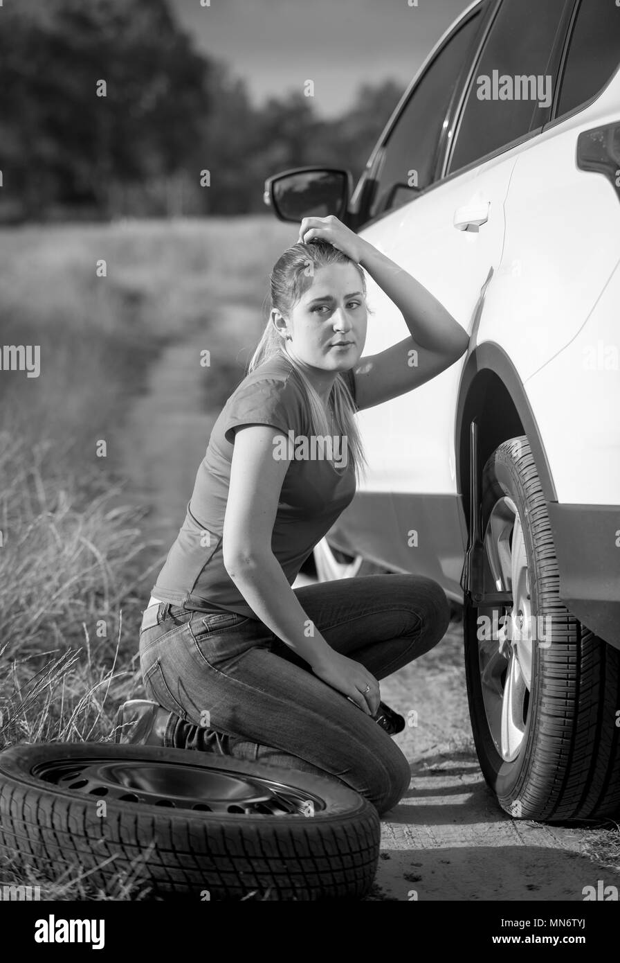 Upset young woman sitting at her car with flat tire in field Stock Photo