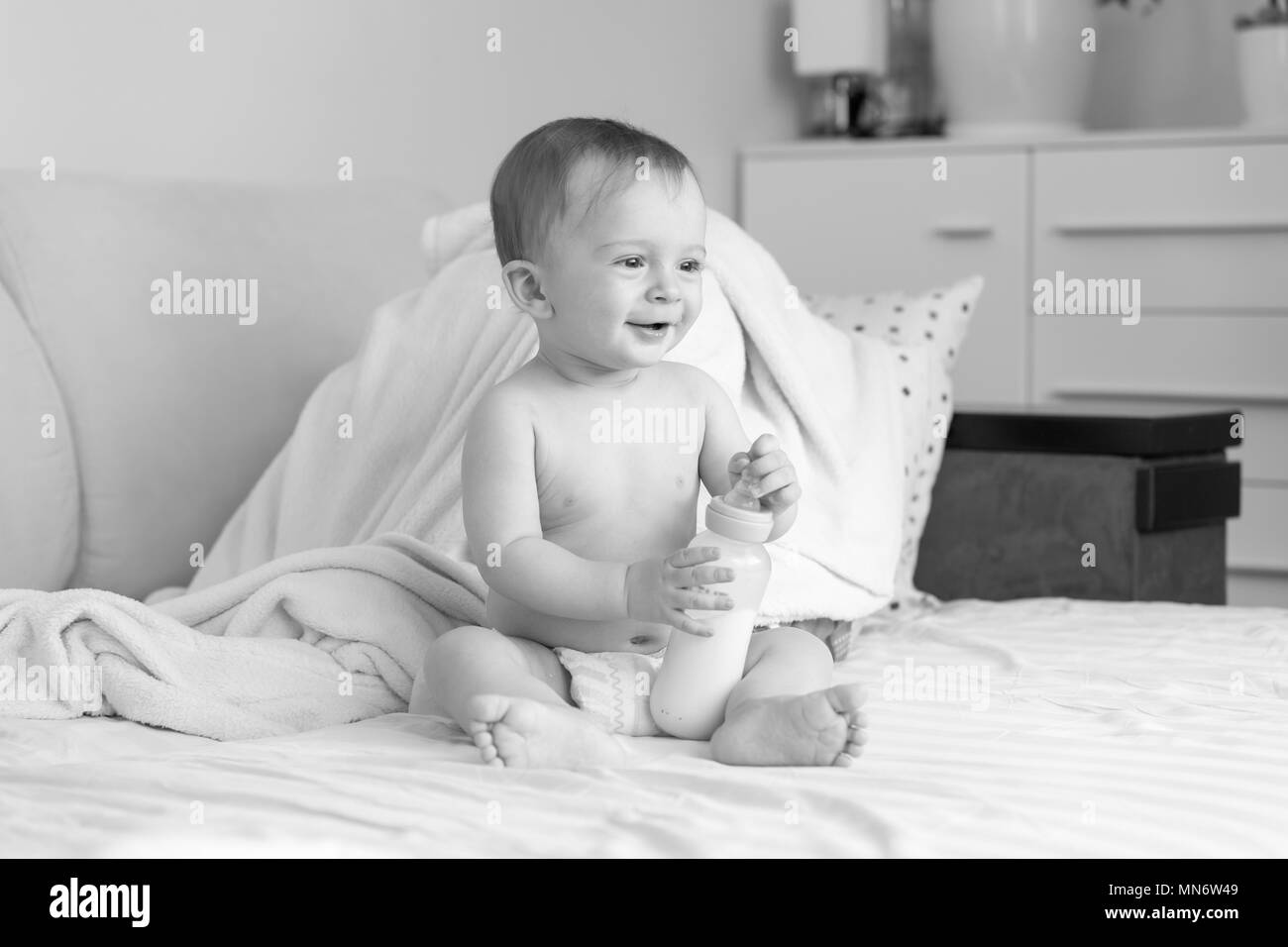 Black and white photo of cute smiling baby boy with bottle of milk sitting on bed Stock Photo