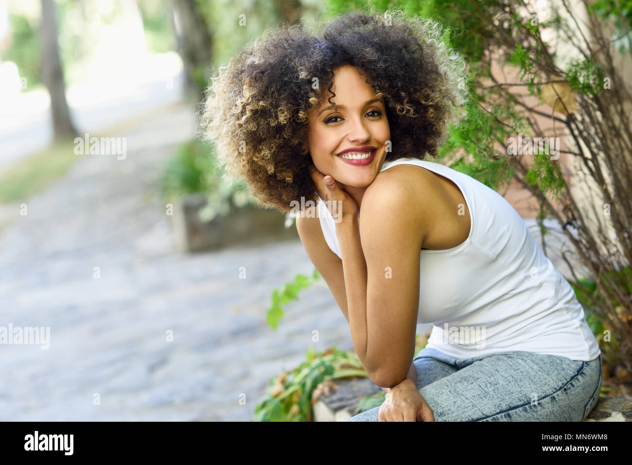 Young Black Woman With Afro Hairstyle Sitting And Smiling In Urban
