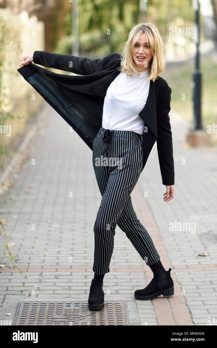 Funny blonde woman smiling in urban background. Young girl wearing black  blazer jacket and striped trousers standing in the street. Pretty female  with straight hair hairstyle and blue eyes Stock Photo -
