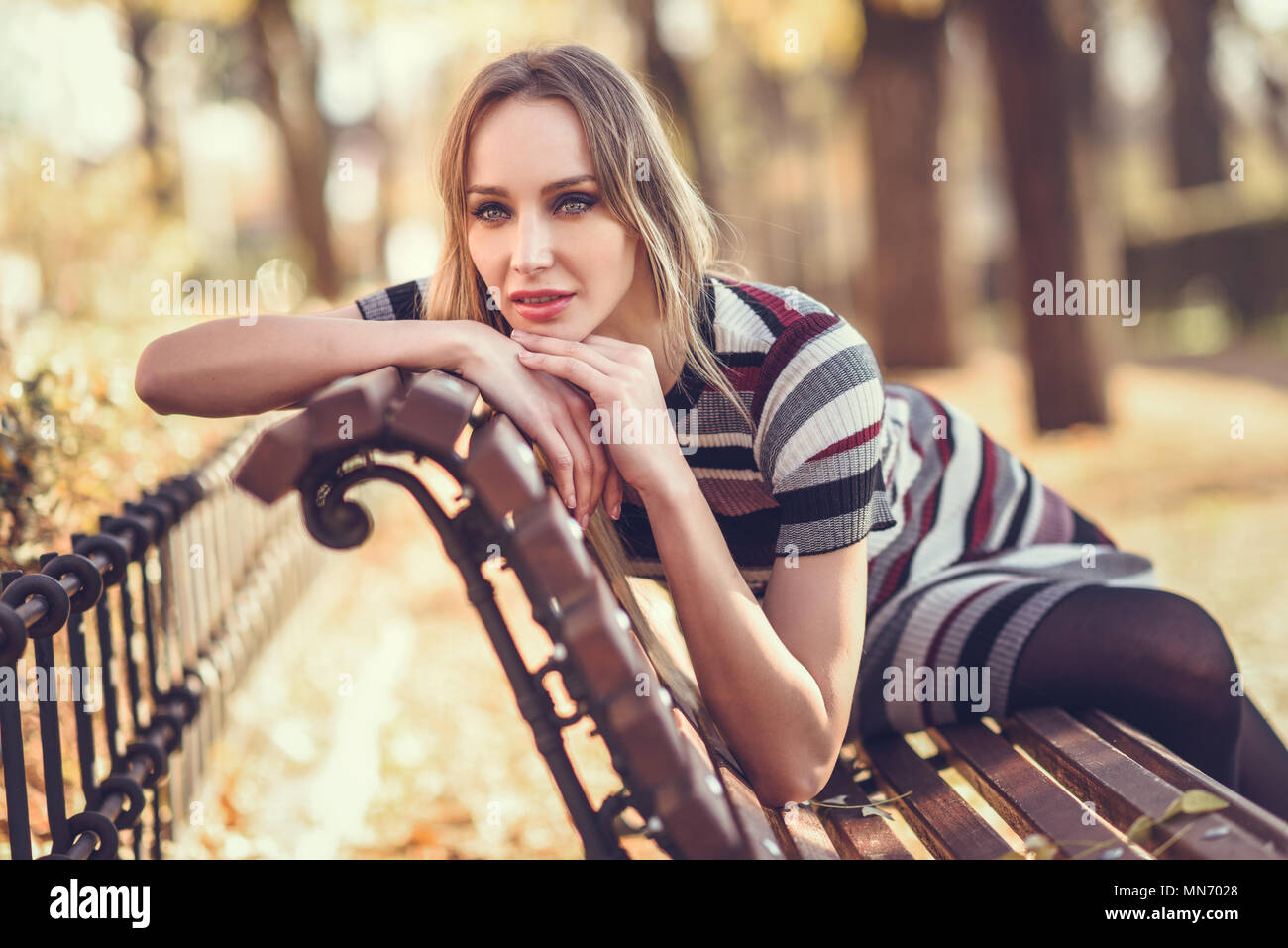 Pensive young blonde woman sitting on a bench in the street of a park ...
