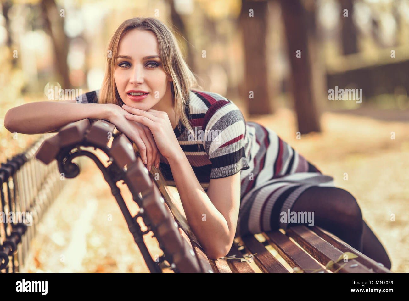 Pensive young blonde woman sitting on a bench in the street of a park ...