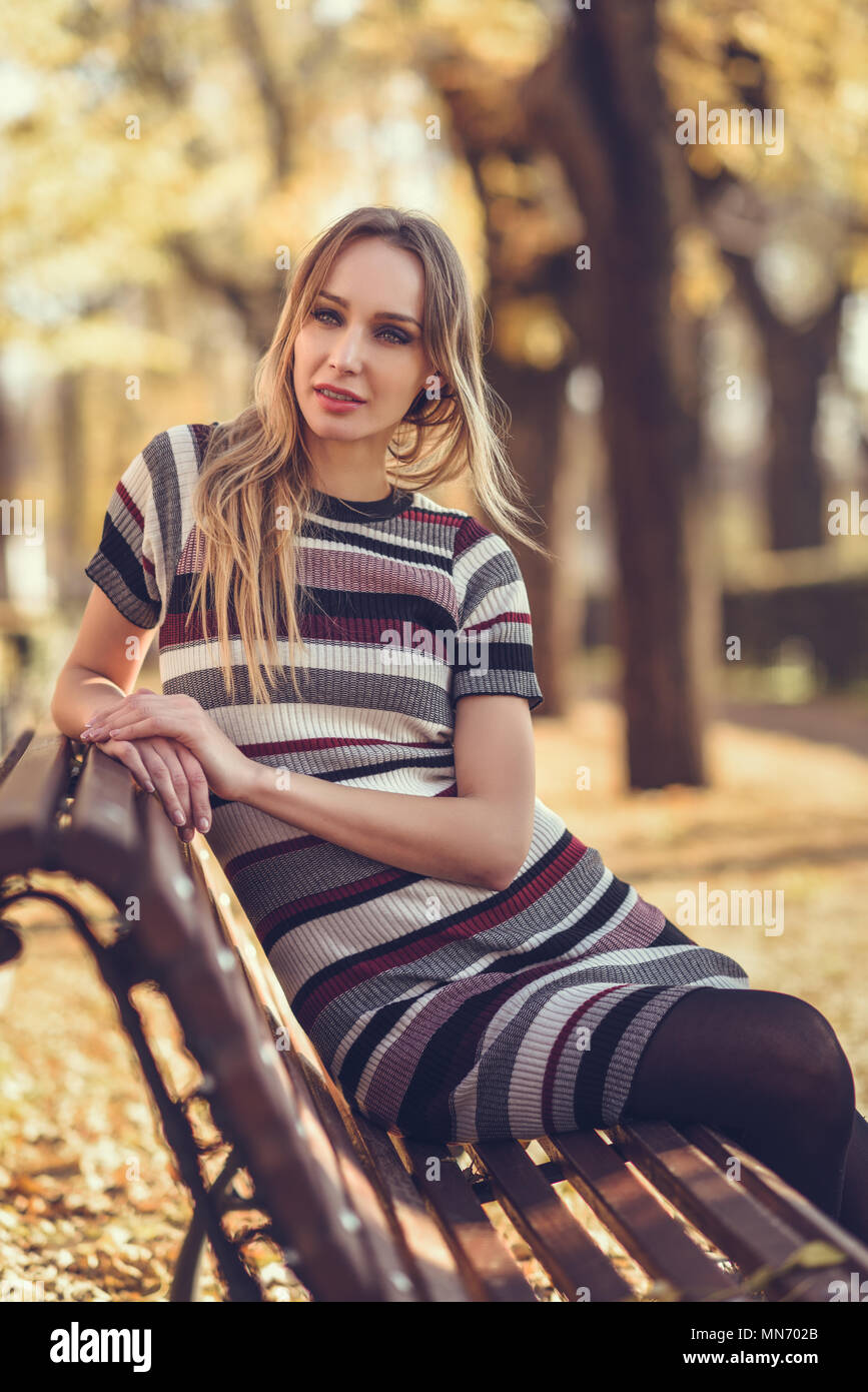 Pensive young blonde woman sitting on a bench in the street of a park ...