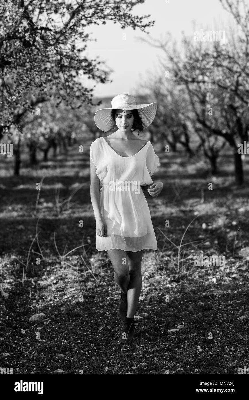 Portrait of young woman in the flowered field in the spring time. Almond flowers blossoms. Girl wearing white dress and pink sun hat Stock Photo