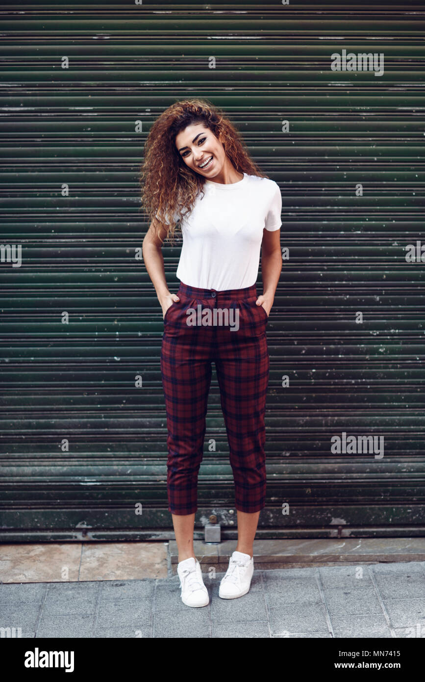 Smiling Young Arabic Woman With Black Curly Hairstyle Arab Girl In Casual Clothes In The Street Happy Female Wearing White T Shirt And Checked Pants Against Urban Blinds Stock Photo Alamy