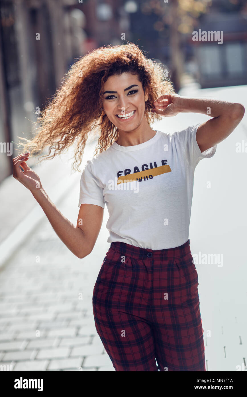 Smiling young arabic woman with black curly hairstyle. Arab girl in casual  clothes in the street. Happy female wearing white t-shirt and checked pants  Stock Photo - Alamy