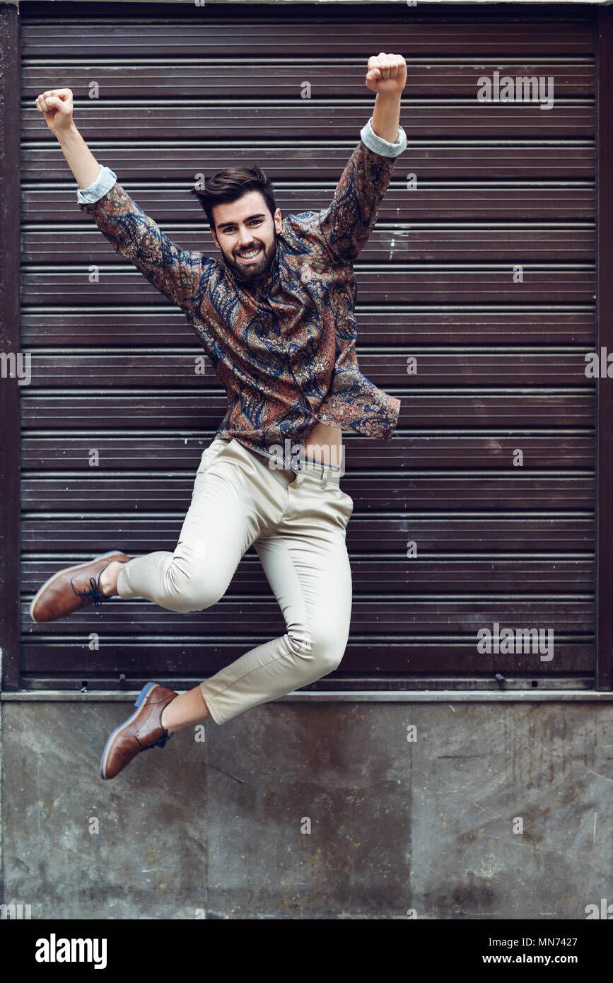 Young bearded man jumping in urban background with open arms wearing casual clothes. Guy with beard and modern hairstyle in the street. Stock Photo