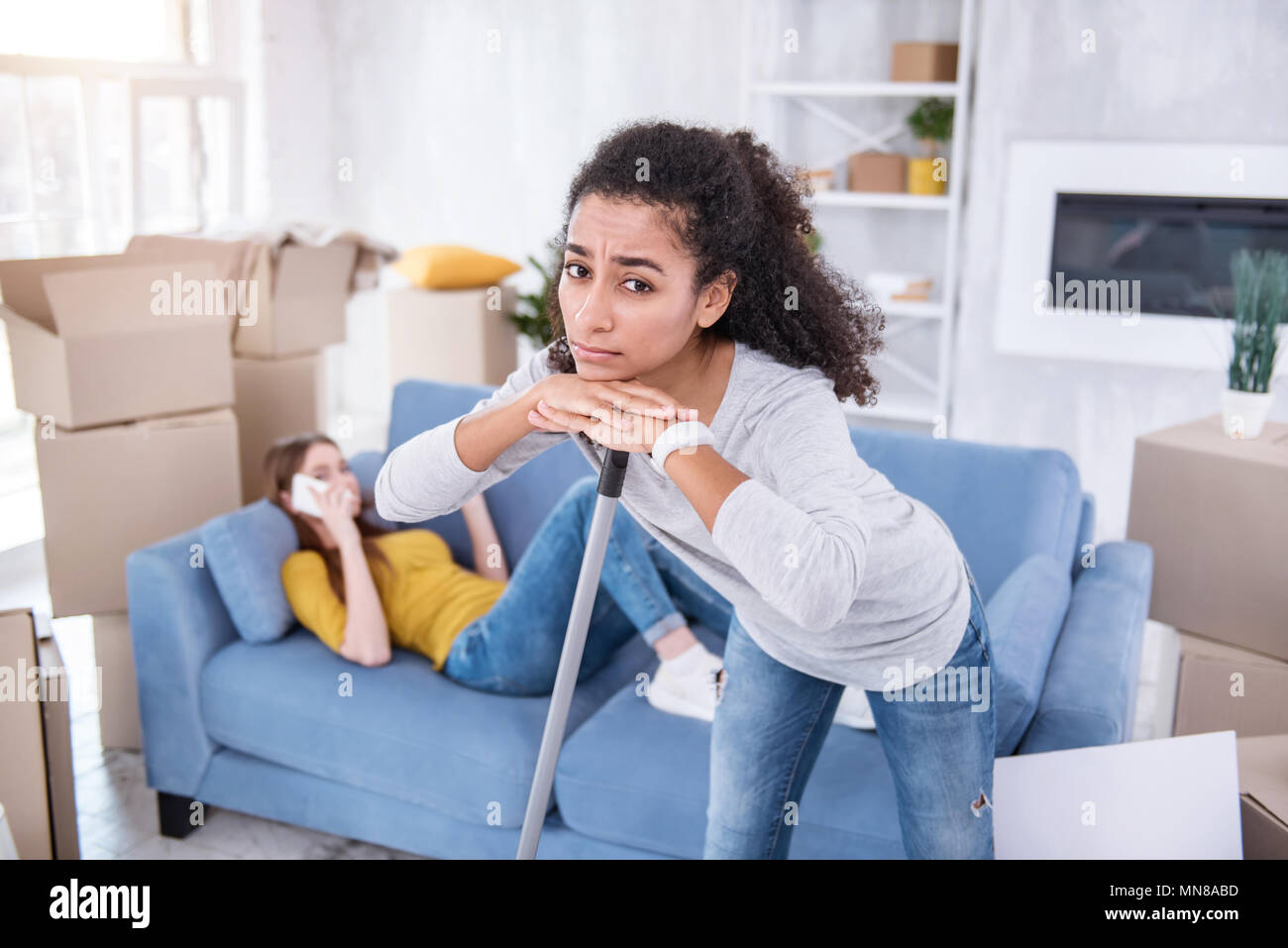 Sad curly girl cleaning apartment while roommate being lazy Stock Photo