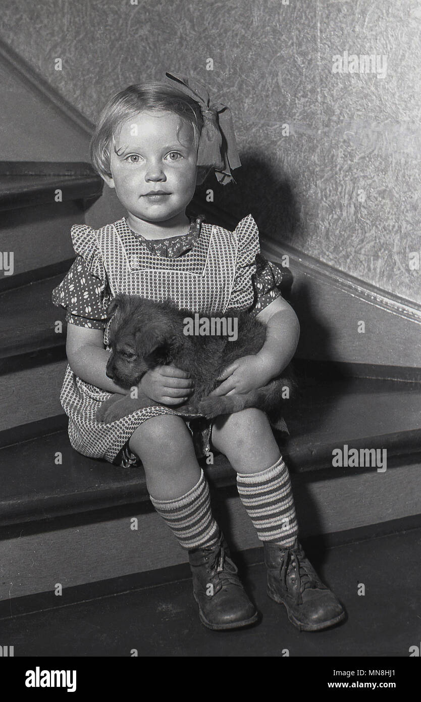 1930s, historical picture showing a sweet little girl seating inside on the bottom of some wooden stairs holding close to her in her lap, her small pet dog. Stock Photo