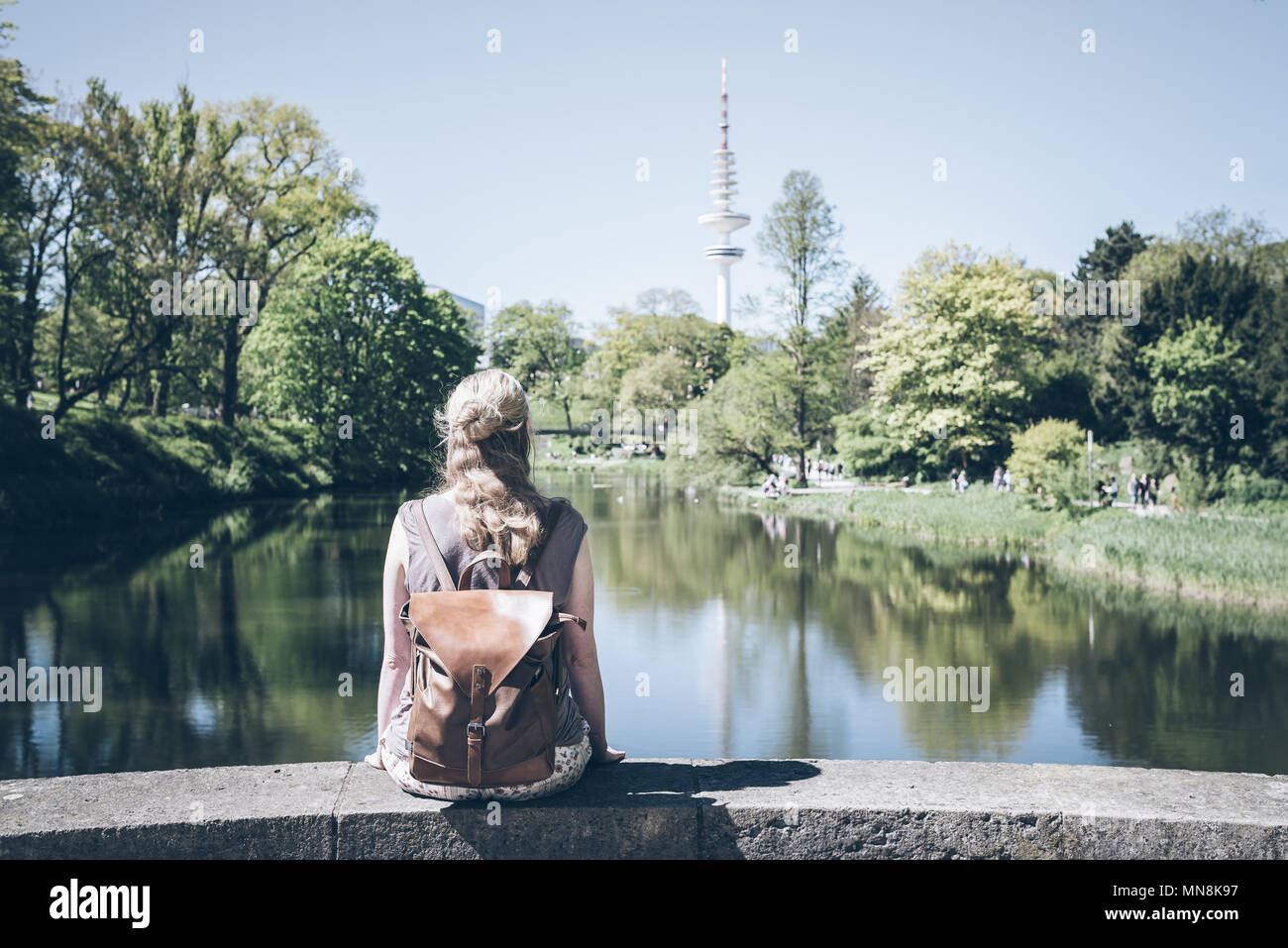 back view of blonde woman sitting on wall by pond in park on sunny day Stock Photo