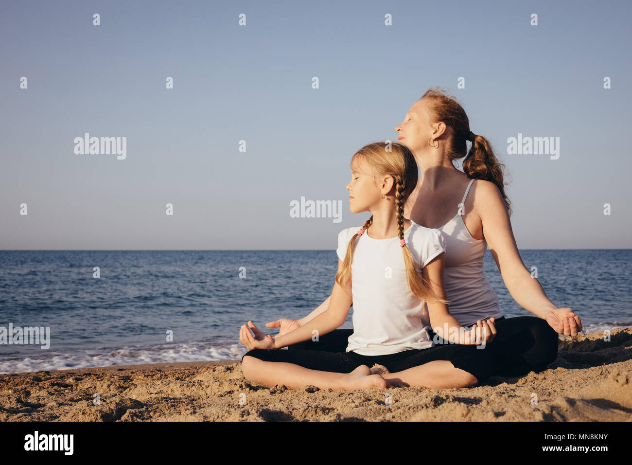Mother and daughter doing yoga exercises  on the beach at the day time. People having fun outdoors. Concept of summer vacation and friendly family. Stock Photo
