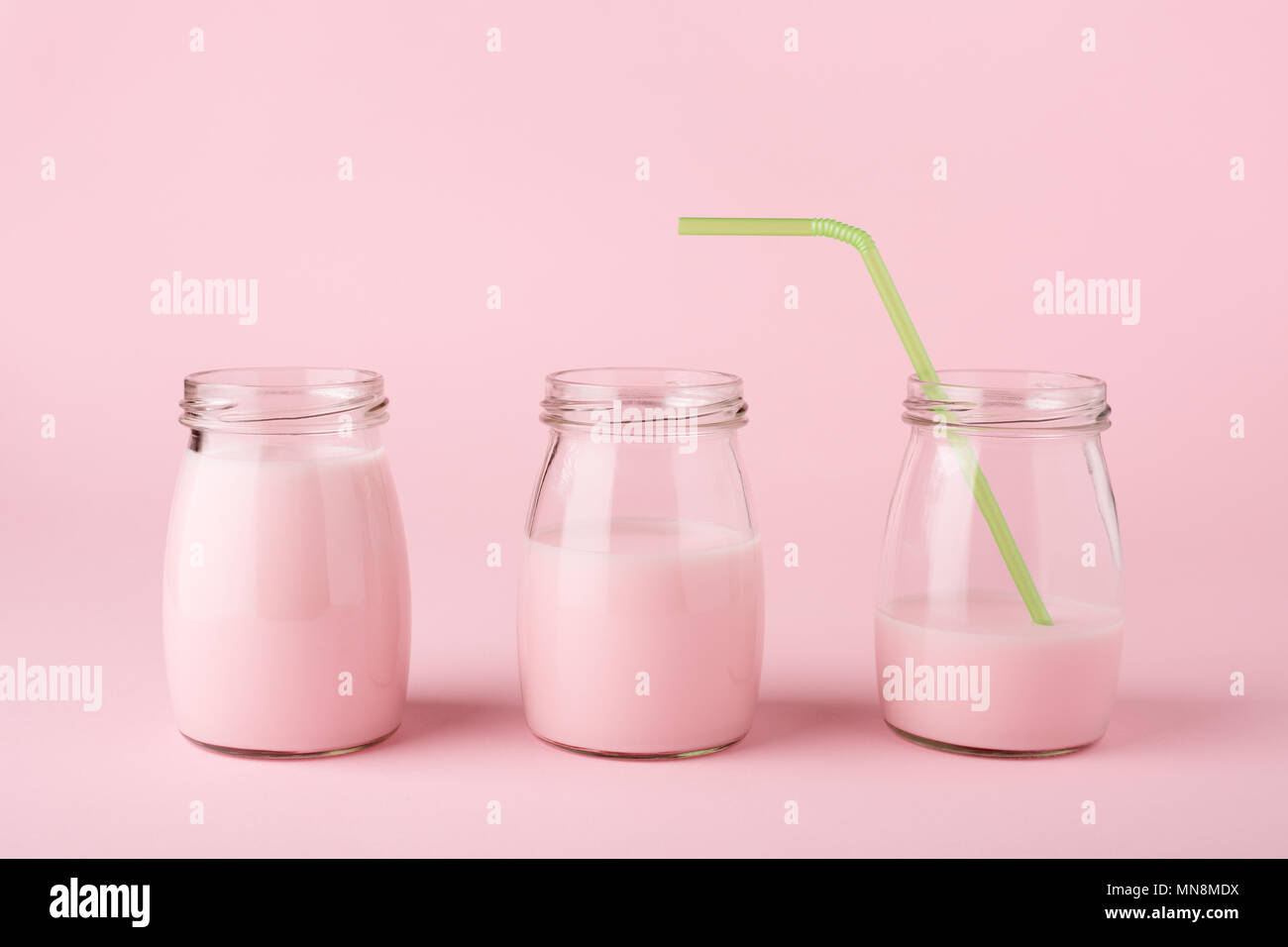 Three glass bottles of fruit yogurt with green straw on the pink background. Minimal style. Playful still life, side view. Healthy drink. Stock Photo