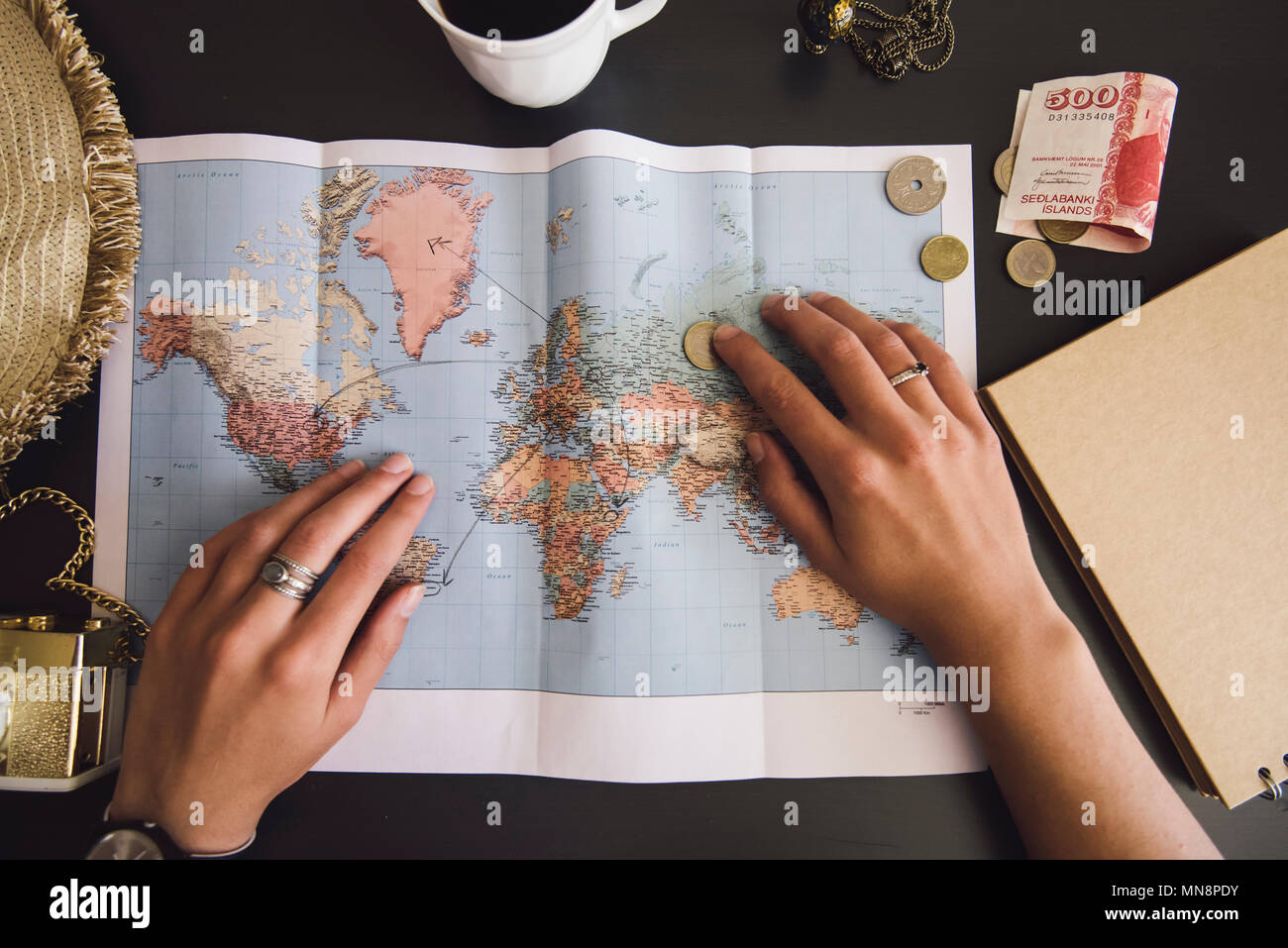 Woman planning her trip. Human hands on the world map with the hat, film camera, some money, notebook from recycled paper and freshly brewed coffee cu Stock Photo