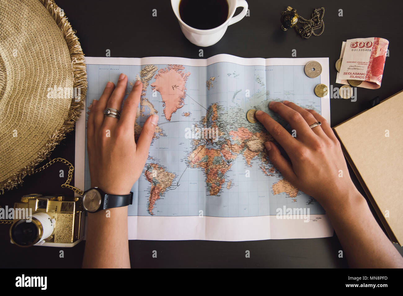 Woman planning her trip. Human hands on the world map with the hat, film camera, some money, notebook from recycled paper and freshly brewed coffee cu Stock Photo