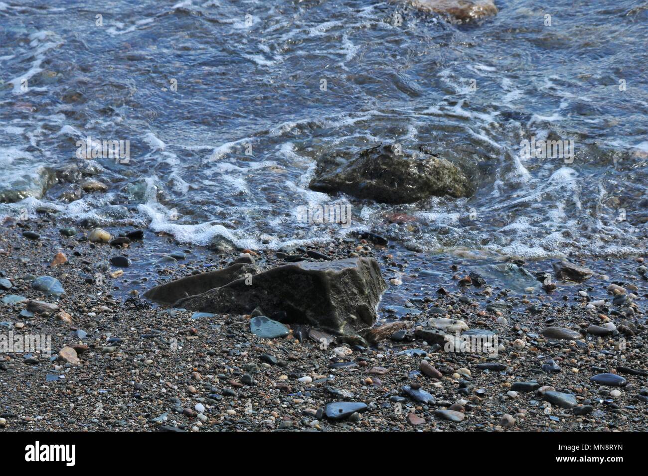 Gentle waves lapping against small rocks on a peddle beach on a sunny day. Stock Photo
