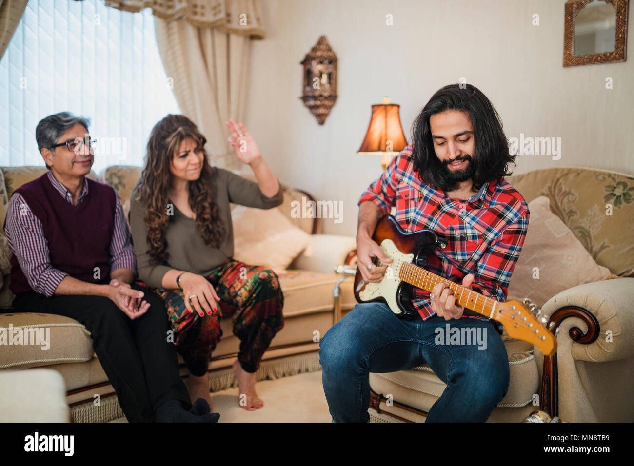 Mid adult man is playing guitar while visiting his parents home. They are sitting on the sofa, dancing and singing along in the background. Stock Photo