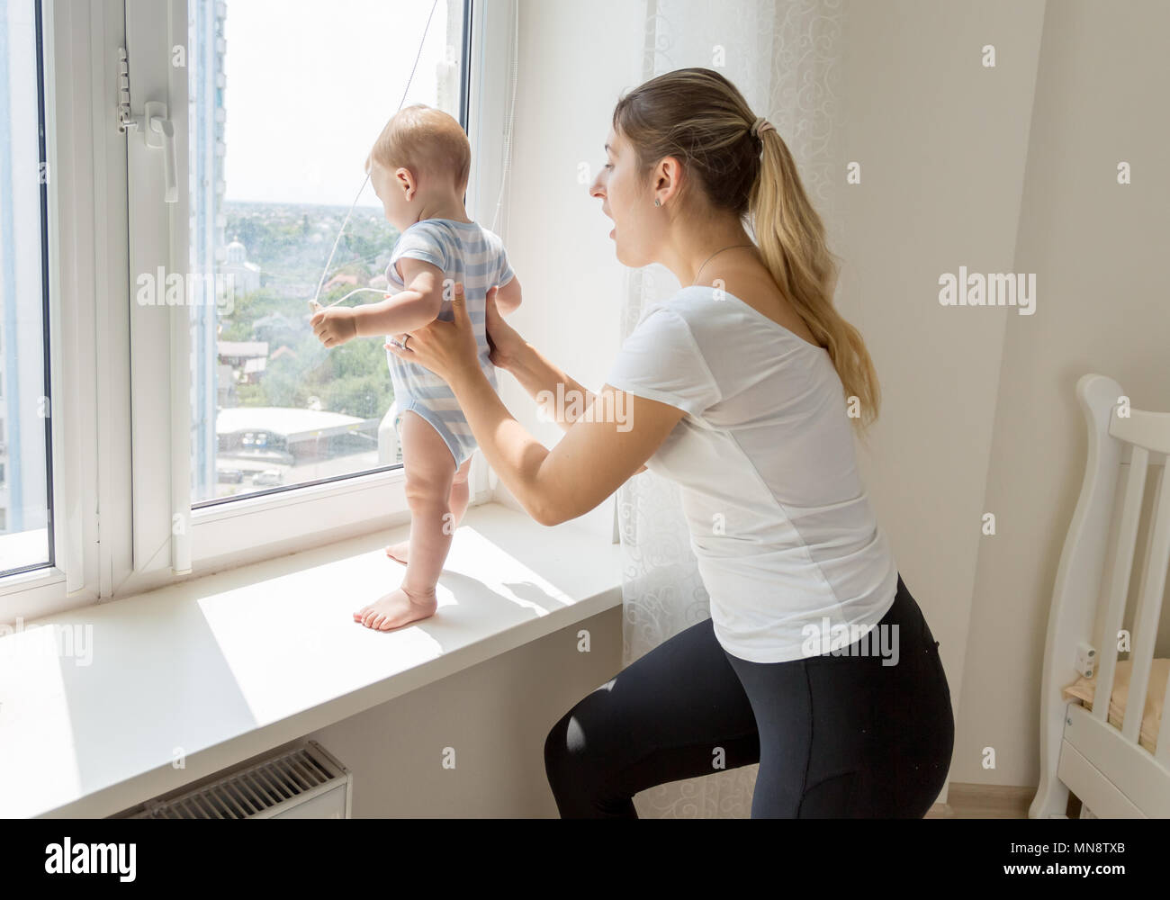 Young mother catching and holding her baby son standing on sindowsil and looking out of the window Stock Photo