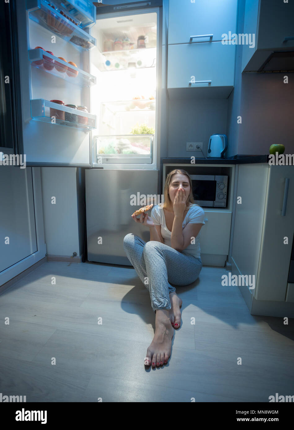 Sleepy hungry woman sitting on kitchen floor at night and holding pizza Stock Photo
